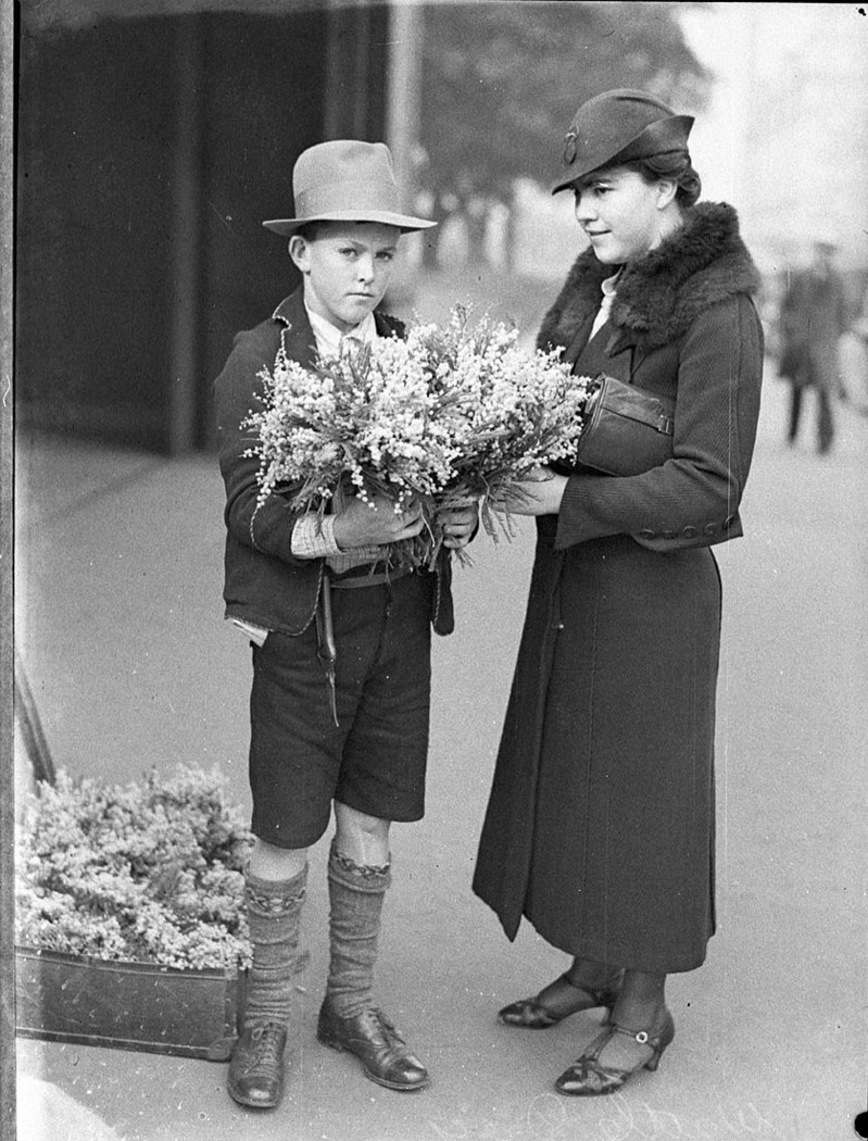 Boy in a hat stands holding bunches of wattle. A woman stands beside him.